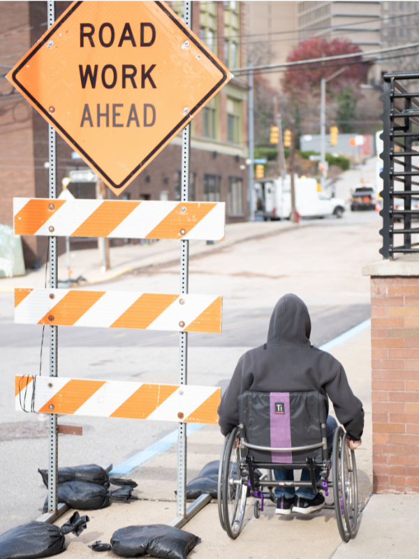 Image of a wheelchair user struggling to navigate around a construction sign on the sidewalk